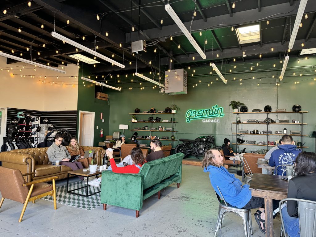 People sit and socialize on couches and at tables in a stylish garage-themed cafe with green walls, string lights, and motorcycle decor. A neon sign reads “Gremlin Garage.”.