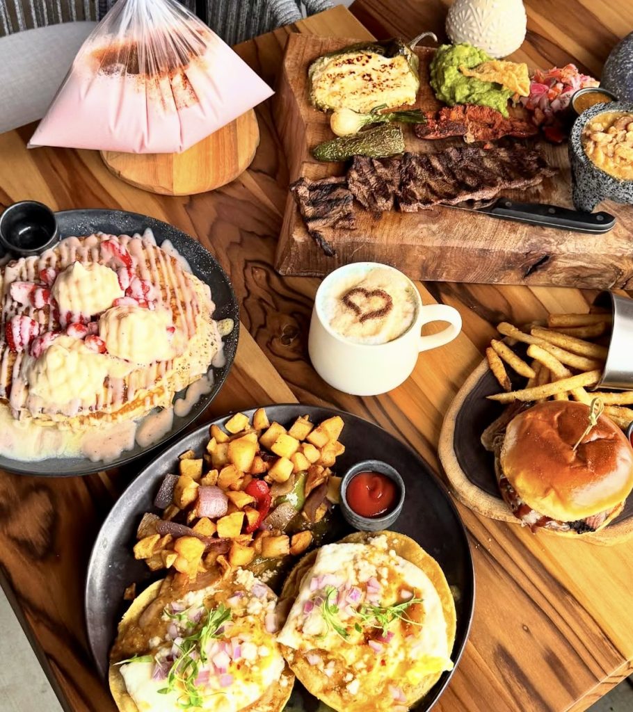 A wooden table topped with various foods: strawberry-topped pancakes, two tacos with potatoes, a burger with fries, grilled meat with guacamole, a cup of coffee with latte art, and a bag of pink drink.