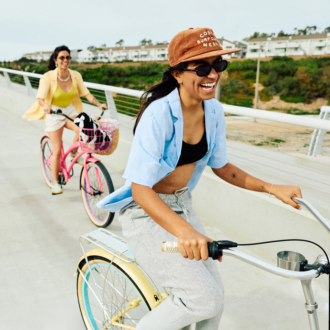 Two women ride bikes on a sunny day along a Costa Mesa path. Both are smiling; the woman in front wears a cap, sunglasses, and a blue shirt, while the woman behind has a yellow shirt and rides a pink bike with a basket. Greenery is visible in the background.