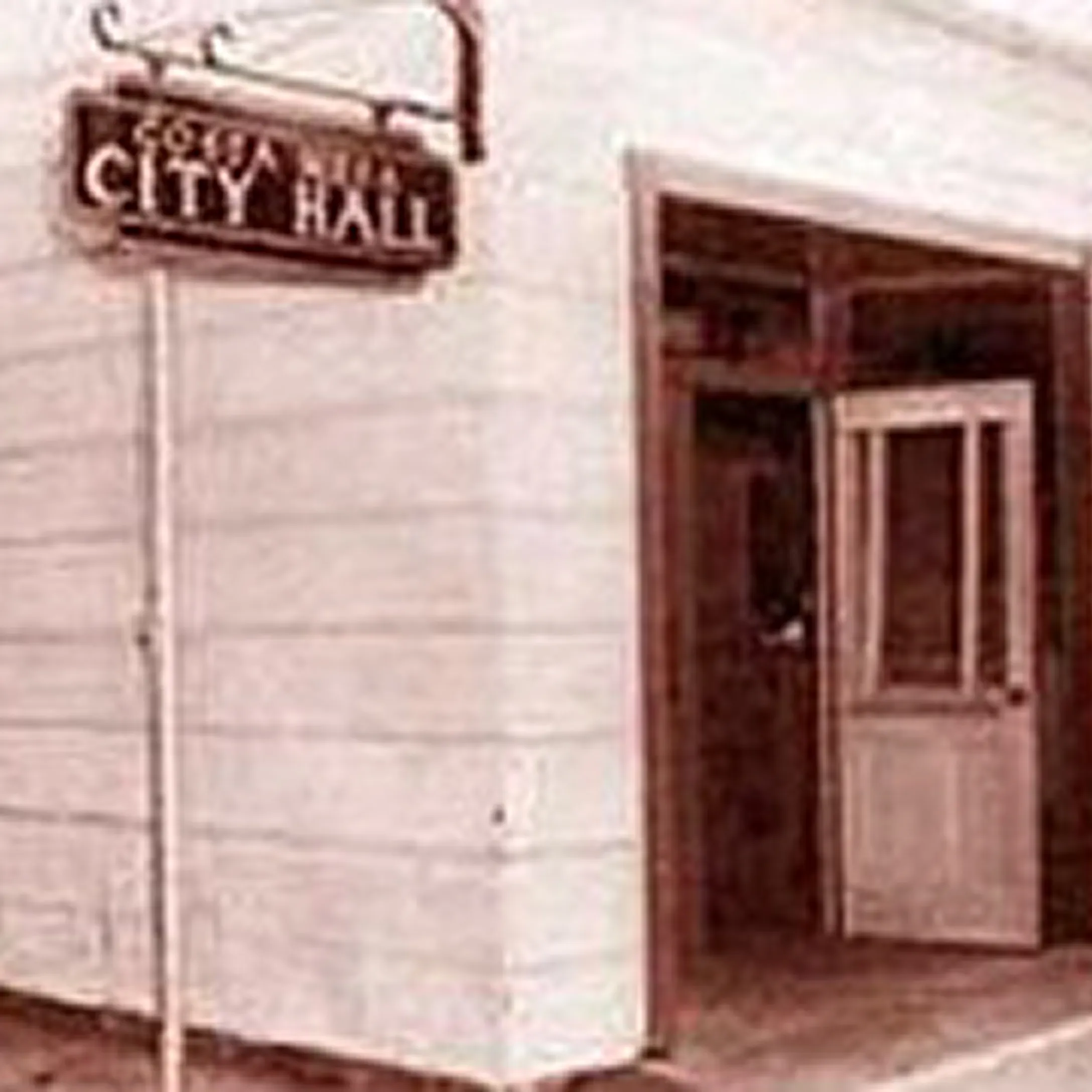 Sepia-toned photo of a building entrance with an open door and a sign above reading CITY HALL. The structure appears old, reflecting the rich history of Costa Mesa, and the sign has some faded, unreadable text above CITY HALL.