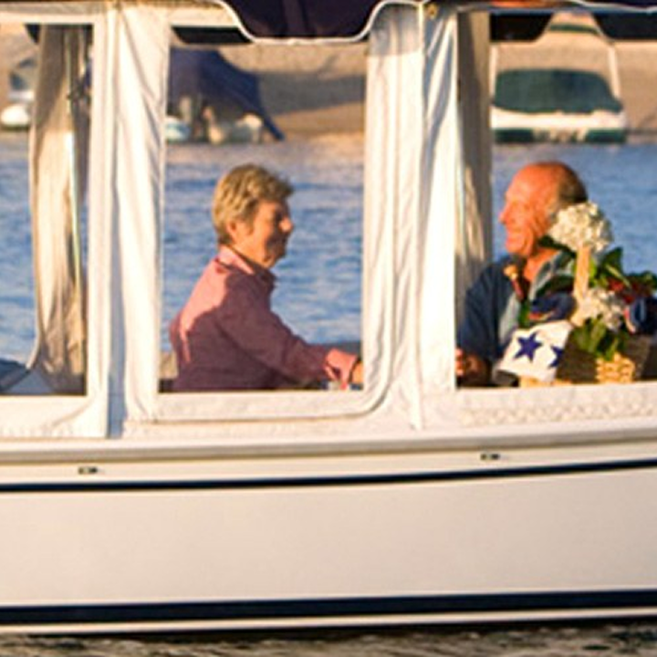Two older adults sit inside a small boat with windows, smiling and talking. There are flowers and water visible in the background.