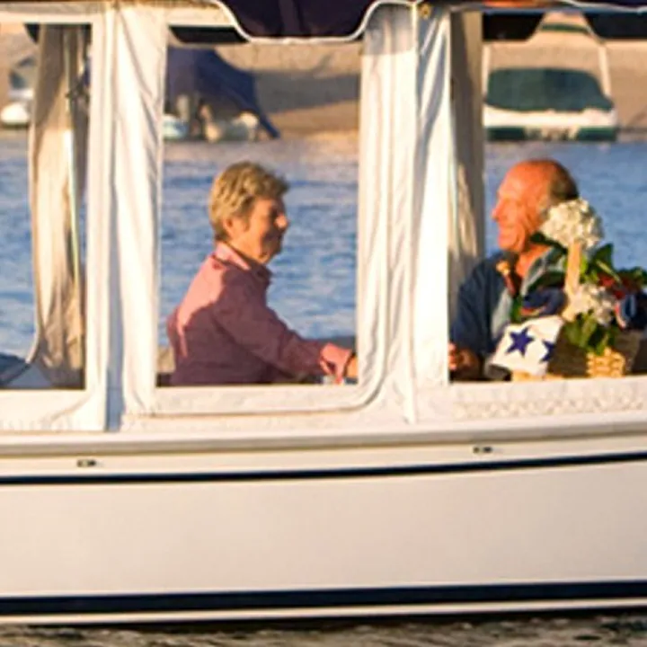 Two older adults sit inside a small boat with windows, smiling and talking. There are flowers and water visible in the background.