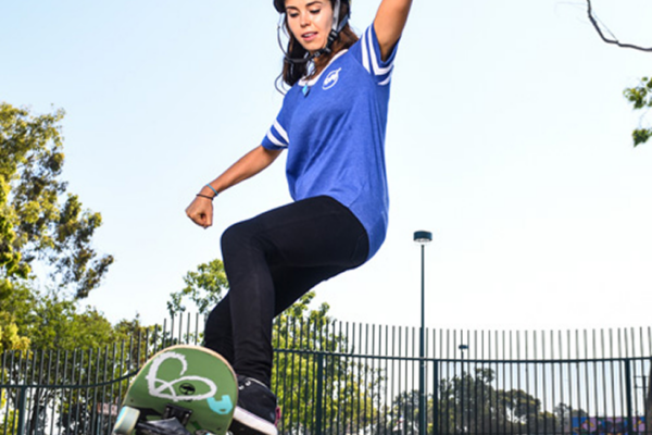 A young woman wearing a helmet and blue shirt is skateboarding at a skate park near beaches near Costa Mesa, performing a trick on the edge of a ramp with trees and a fence in the background.