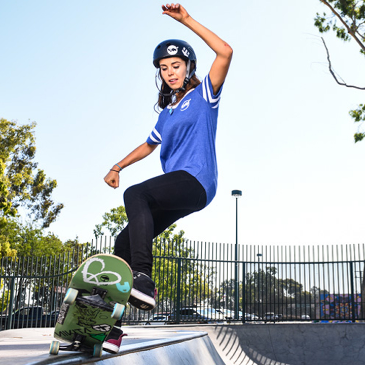 A young woman wearing a helmet and blue shirt is skateboarding at a skate park near beaches near Costa Mesa, performing a trick on the edge of a ramp with trees and a fence in the background.