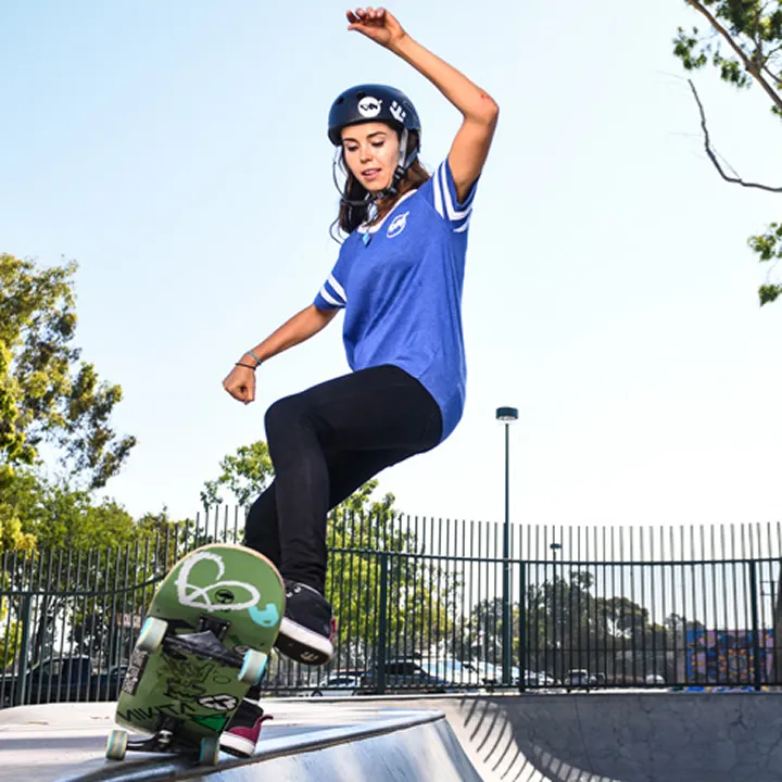 A young woman wearing a helmet and blue shirt is skateboarding at a skate park near beaches near Costa Mesa, performing a trick on the edge of a ramp with trees and a fence in the background.