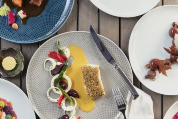 An overhead view of a wooden table set with plates of gourmet dishes reminiscent of Michelin star restaurants in Orange County, featuring fish with sauce, vibrant salads, a dessert, elegant garnishes, and neatly placed cutlery and napkin.