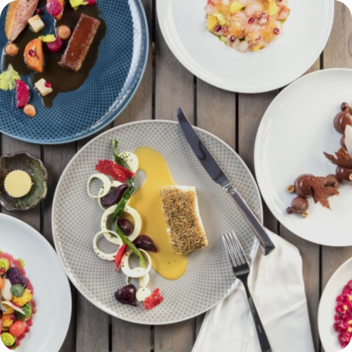 An overhead view of a wooden table set with plates of gourmet dishes reminiscent of Michelin star restaurants in Orange County, featuring fish with sauce, vibrant salads, a dessert, elegant garnishes, and neatly placed cutlery and napkin.