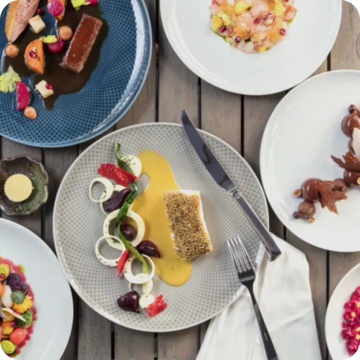 An overhead view of a wooden table set with plates of gourmet dishes reminiscent of Michelin star restaurants in Orange County, featuring fish with sauce, vibrant salads, a dessert, elegant garnishes, and neatly placed cutlery and napkin.