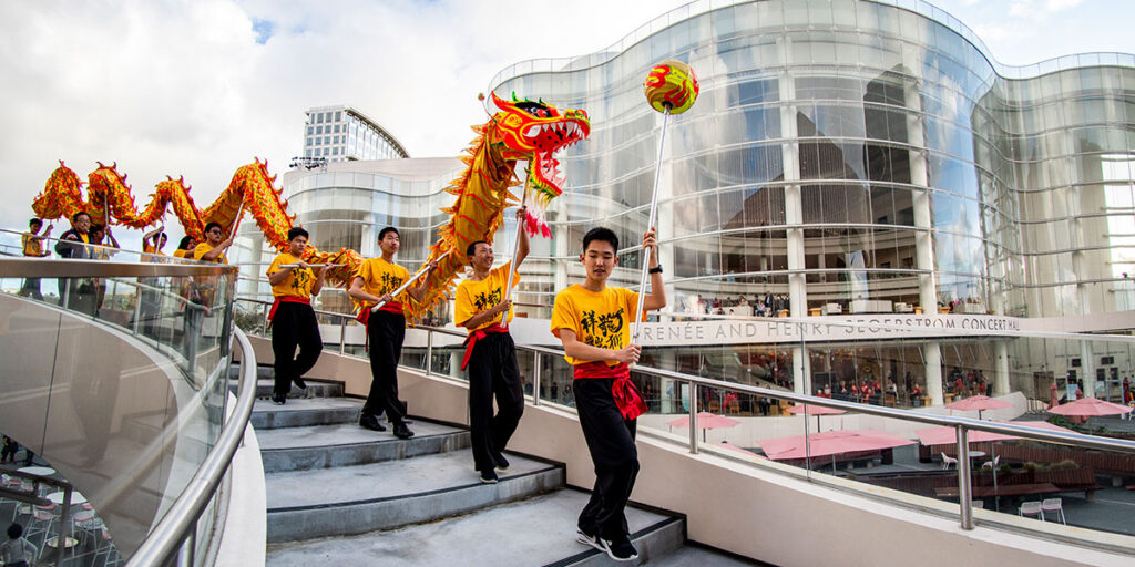 A group performs a traditional dragon dance in yellow shirts on an outdoor staircase, with a modern glass building and people in the background—an eye-catching scene perfect for any Costa Mesa travel blog.