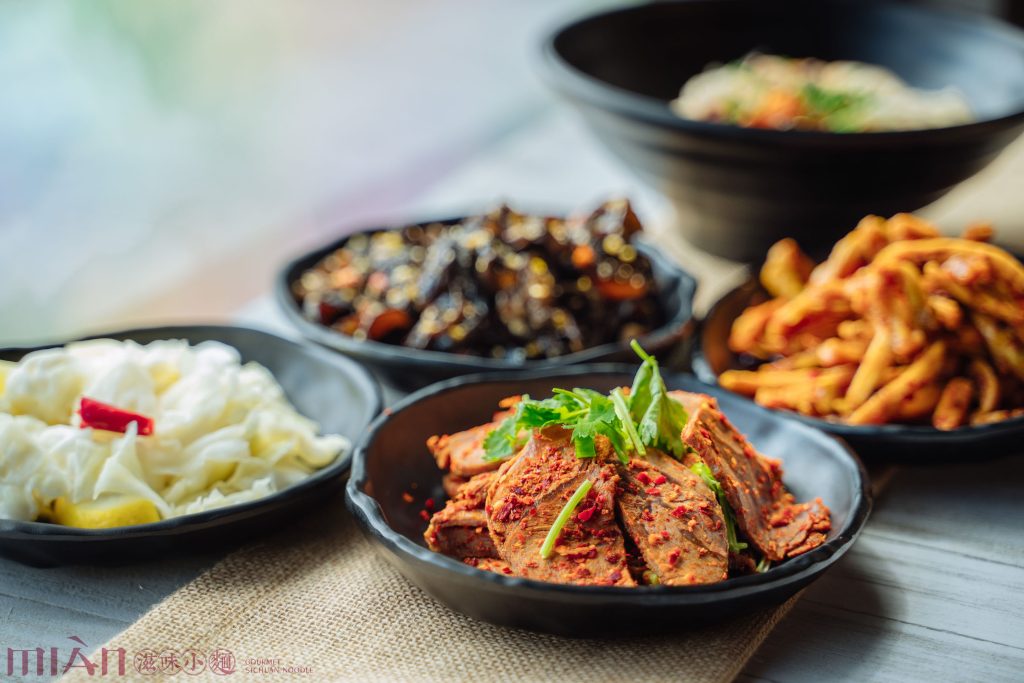 A close-up of several bowls filled with various Asian side dishes, including sliced tofu with red chili flakes, pickled vegetables, and other assorted items, arranged on a table.