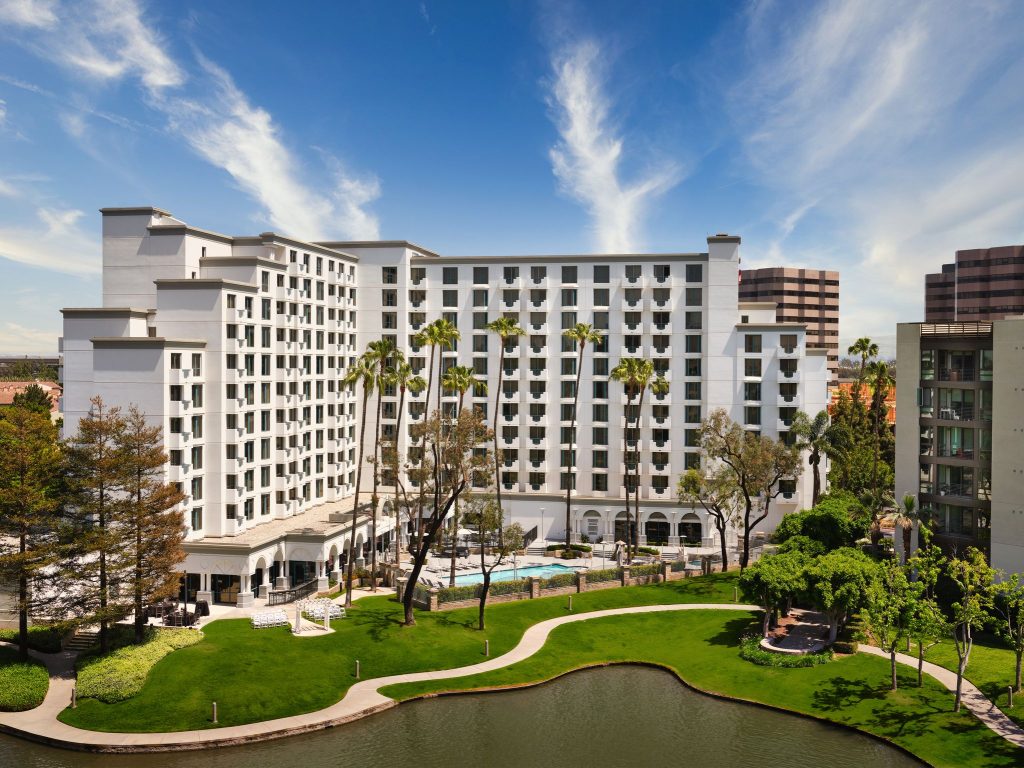 A large white hotel with balconies stands behind a landscaped lawn, palm trees, and a small pond, with a swimming pool visible in the center on a sunny day with blue sky and wispy clouds.