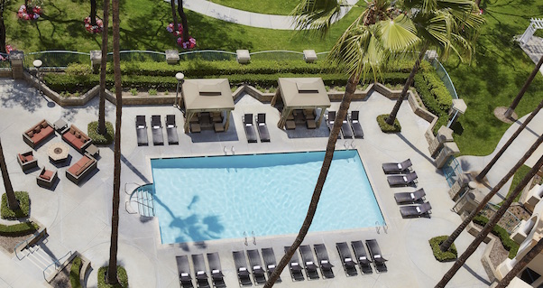 Aerial view of a rectangular outdoor swimming pool surrounded by lounge chairs, cabanas, palm trees, and seating areas, with manicured green lawns and flower beds in the background.