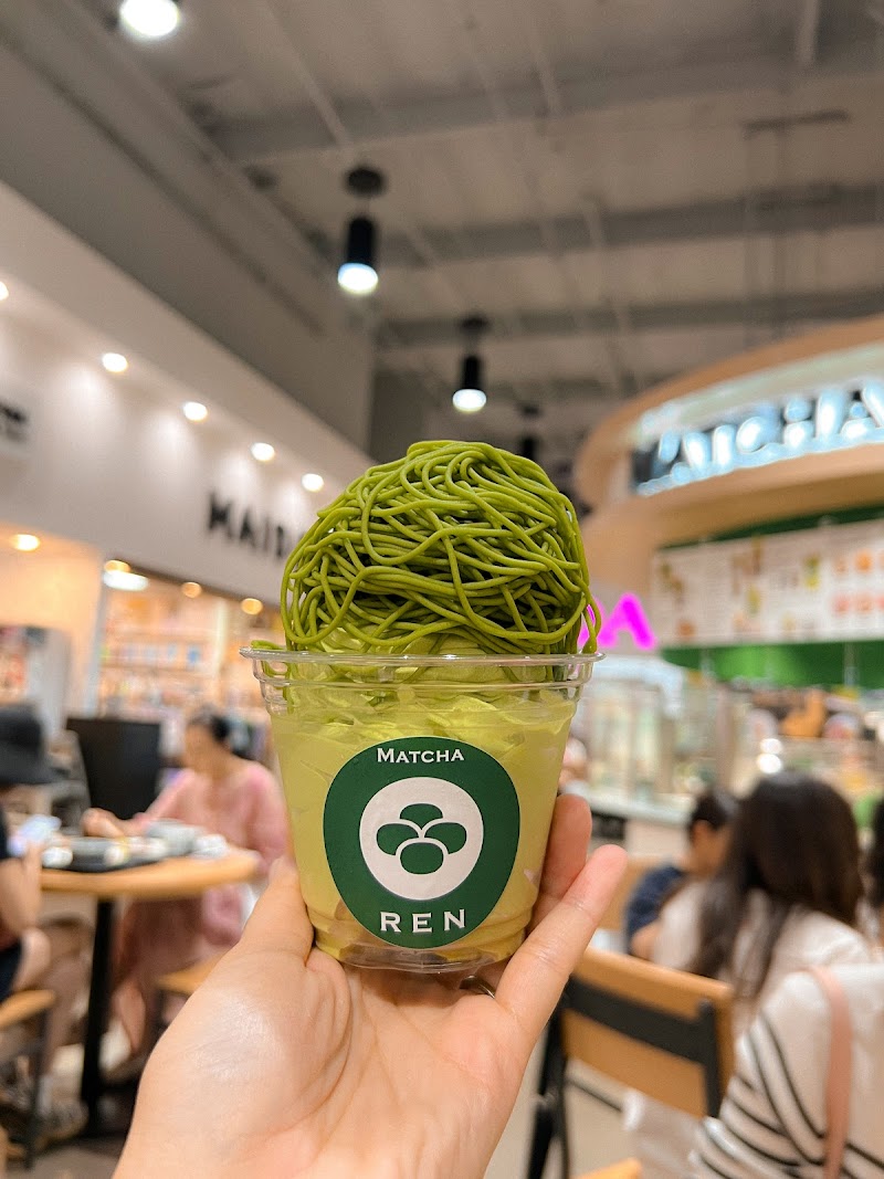 A hand holds a cup of matcha dessert with green matcha noodles on top in a brightly lit café, reminiscent of the vibe at Silver Trumpet Restaurant Costa Mesa, as people sit and talk in the background. The cup is labeled Matcha REN.