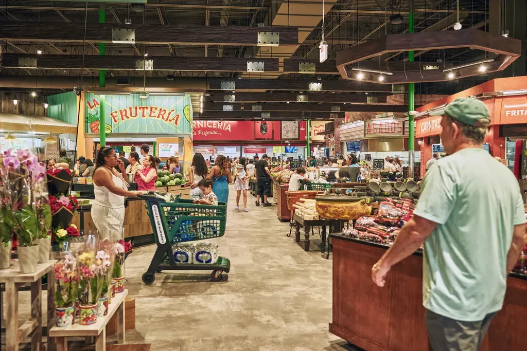 Shoppers browse fresh produce and goods in a busy, vibrant supermarket with signs in Spanish, carts, flower displays, and various market stalls visible throughout the spacious, well-lit store.