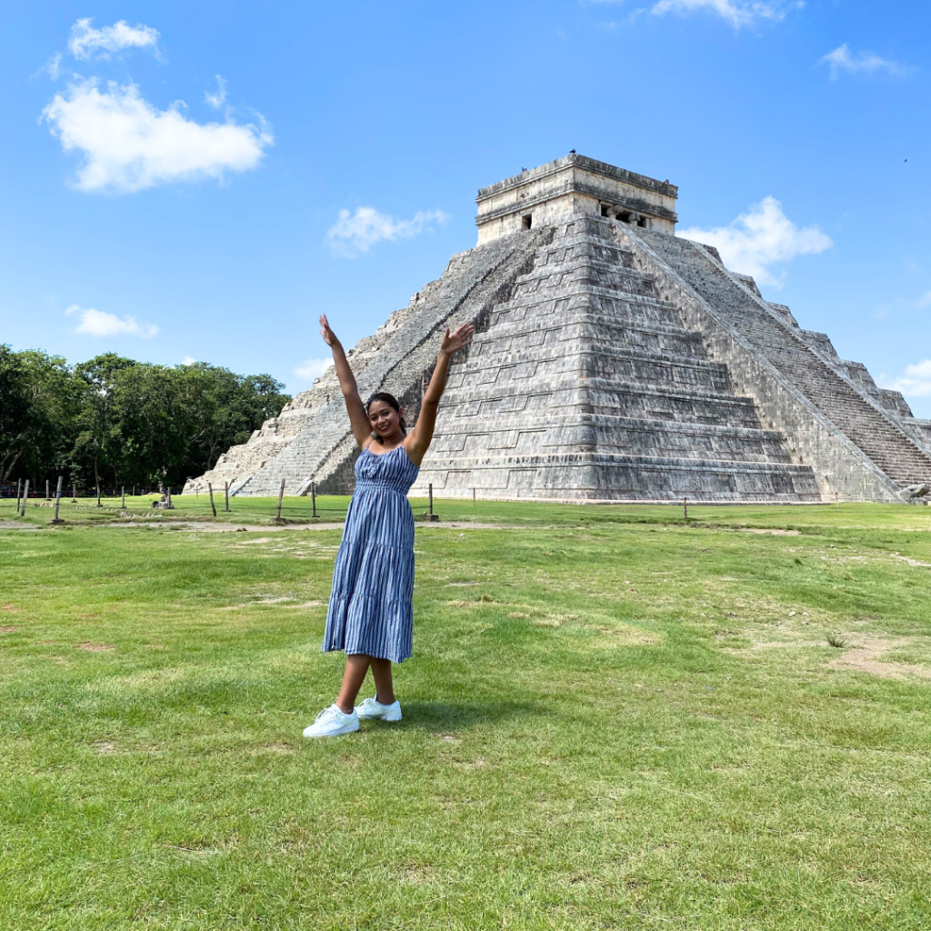A person in a striped dress stands on grass with arms raised, smiling in front of the ancient stone pyramid of Chichen Itza under a bright blue sky with a few clouds.
