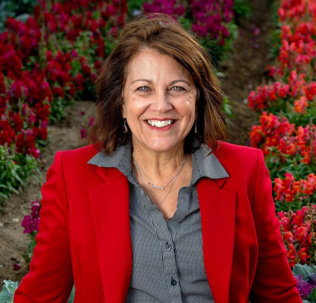 A woman with shoulder-length brown hair wearing a red blazer and patterned shirt smiles while standing in a colorful flower garden with red and pink blooms.