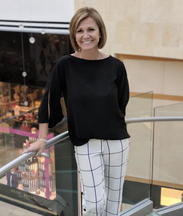 A woman with short light brown hair, wearing a black top and white pants with a black grid pattern, stands smiling on a glass-railed staircase in a shopping mall.