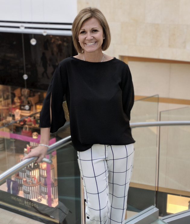 A woman with short hair smiles while standing on a glass-railed staircase. She is wearing a black top and white pants with a grid pattern. A colorful store display is visible in the background.