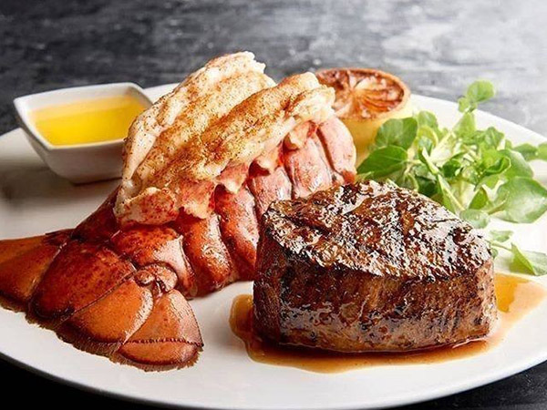 A plate with a grilled lobster tail, a seared steak filet, a small dish of melted butter, a slice of grilled lemon, and a garnish of fresh greens.