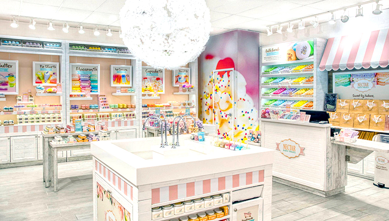 A bright, colorful shop displays bath products like soap bars and bath bombs on shelves. The decor is pastel with candy-themed accents, white furniture, and a large round light fixture hanging from the ceiling.