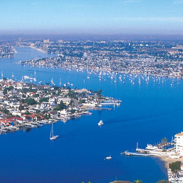 Aerial view of a coastal city near Costa Mesa, featuring a wide blue bay dotted with sailboats and yachts, and densely packed houses and buildings lining the shoreline under a clear sky.