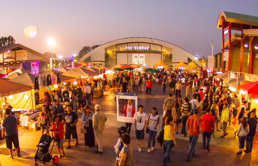 A large outdoor market at sunset in Costa Mesa buzzes with people exploring colorful vendor tents and bright lights, while The Hangar rises in the background—a lively, festive scene perfect for any Costa Mesa travel blog.
