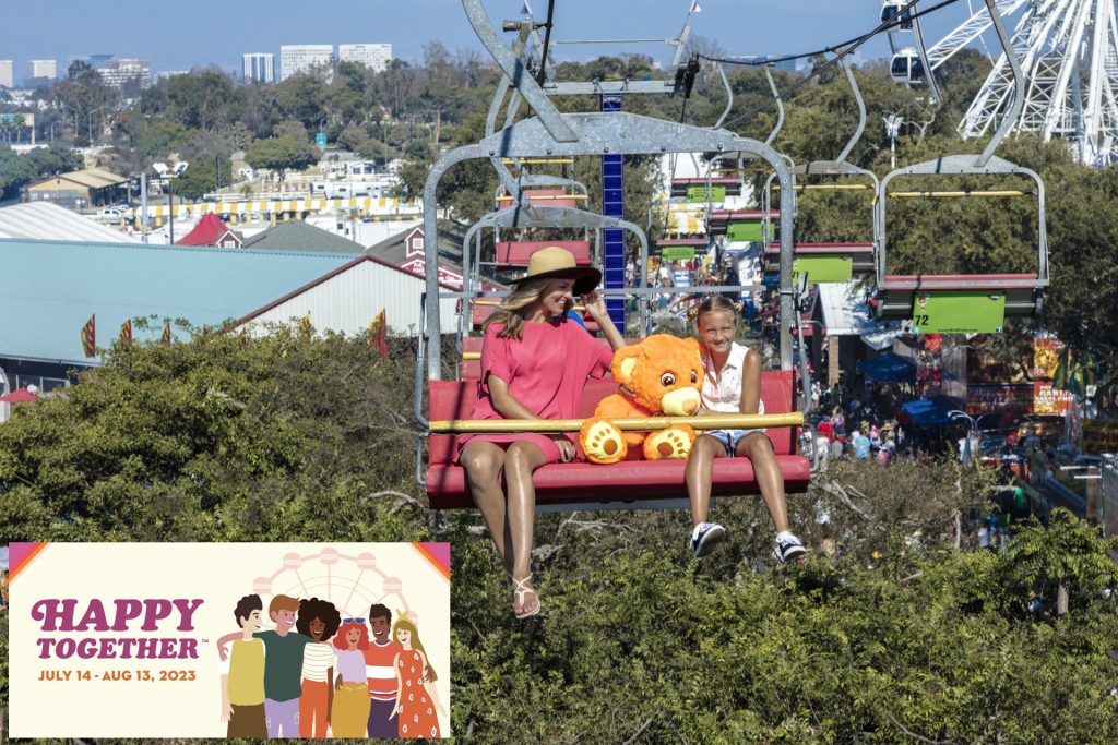 A woman and a child holding a large teddy bear ride a chairlift over a fairground. An illustrated banner in the corner reads Happy Together, July 14 - Aug 13, 2023 with smiling people and a Ferris wheel.