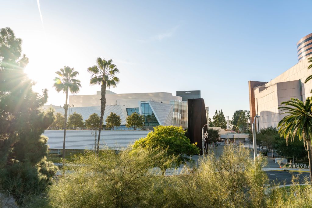 A modern building with angular white architecture is surrounded by palm trees and greenery under a clear blue sky, with sunlight streaming from the left. Urban buildings are visible in the background.