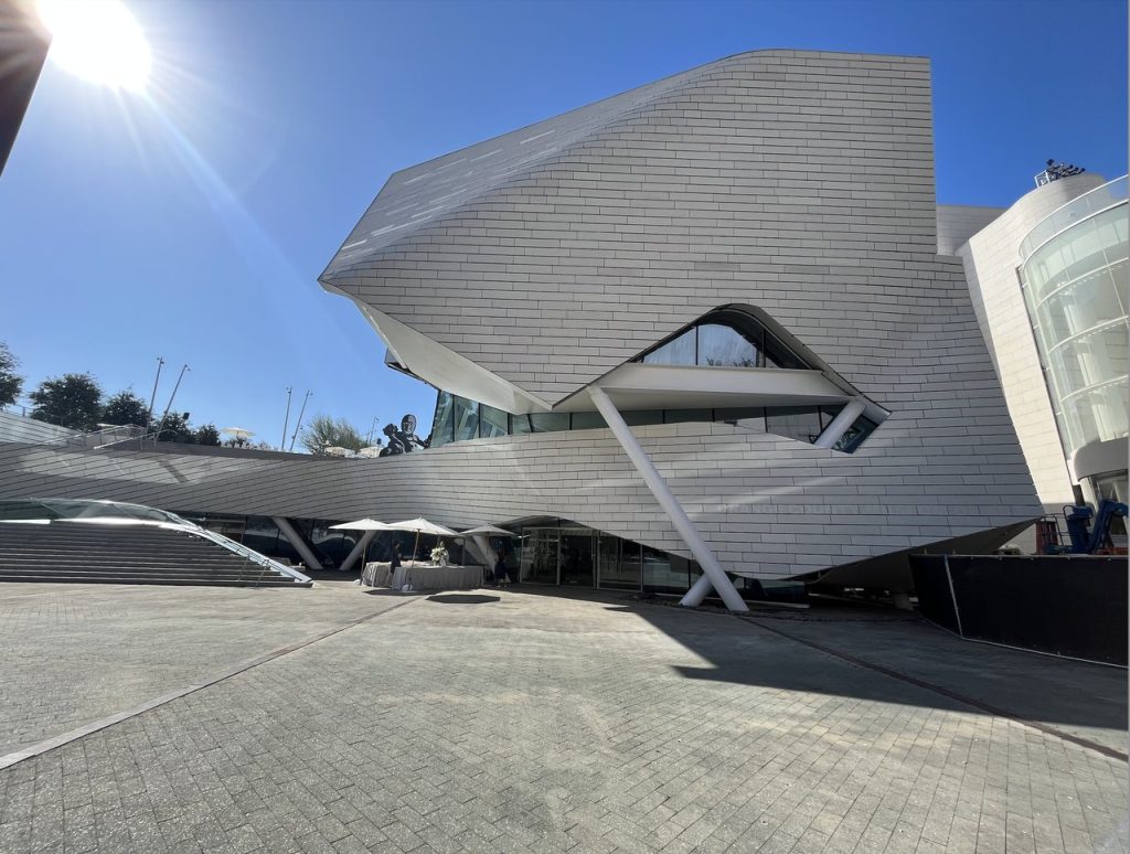 A modern building with angular, futuristic white panels and large windows, bathed in bright sunlight with a clear blue sky in the background. The structure features unique shapes and slanted support beams.