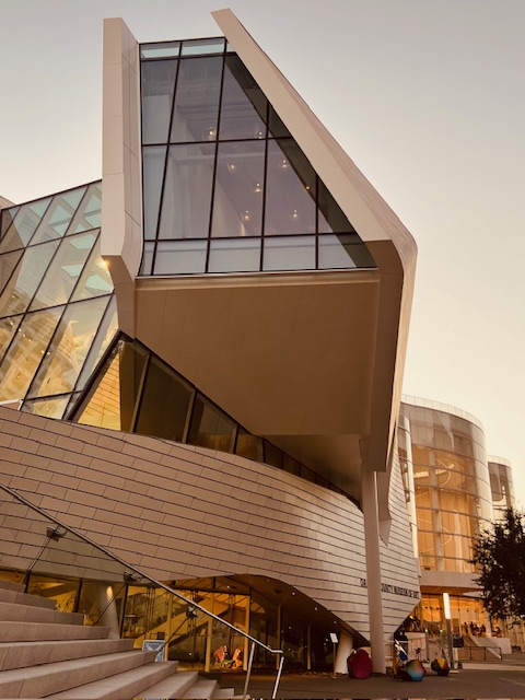 A modern building with sharp angles, large glass windows, and cream-colored exterior walls, photographed at sunset. The architecture features an overhanging section supported by pillars and contemporary design elements.