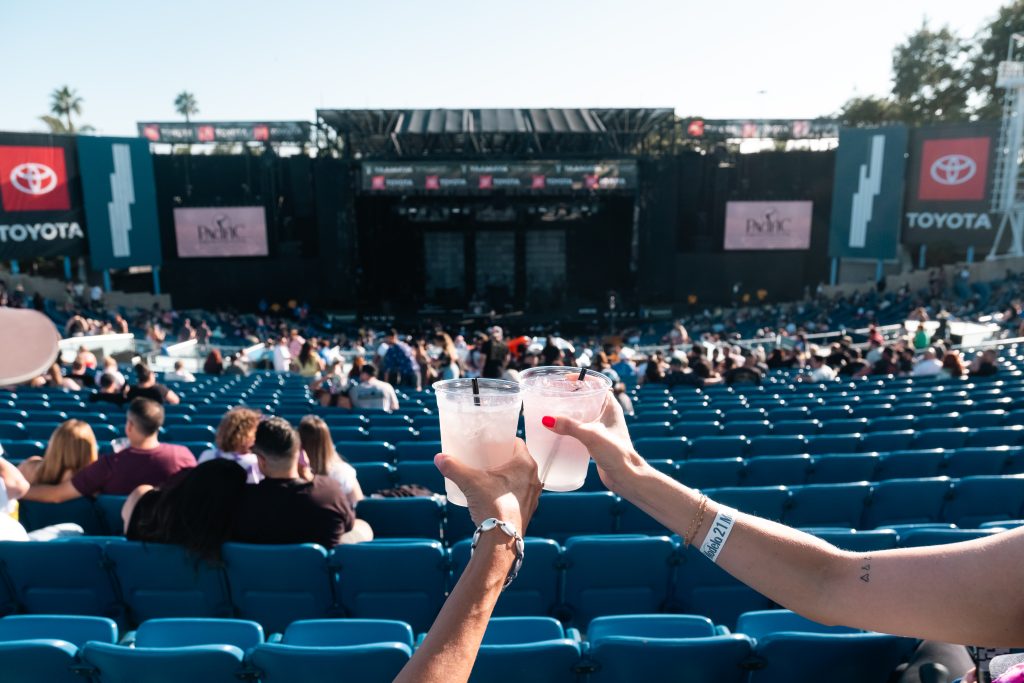 Two people hold up drinks in plastic cups, toasting at an outdoor concert venue with blue seats and a large stage in the background. The audience is filling in, and branded Toyota signs are visible on stage sides.