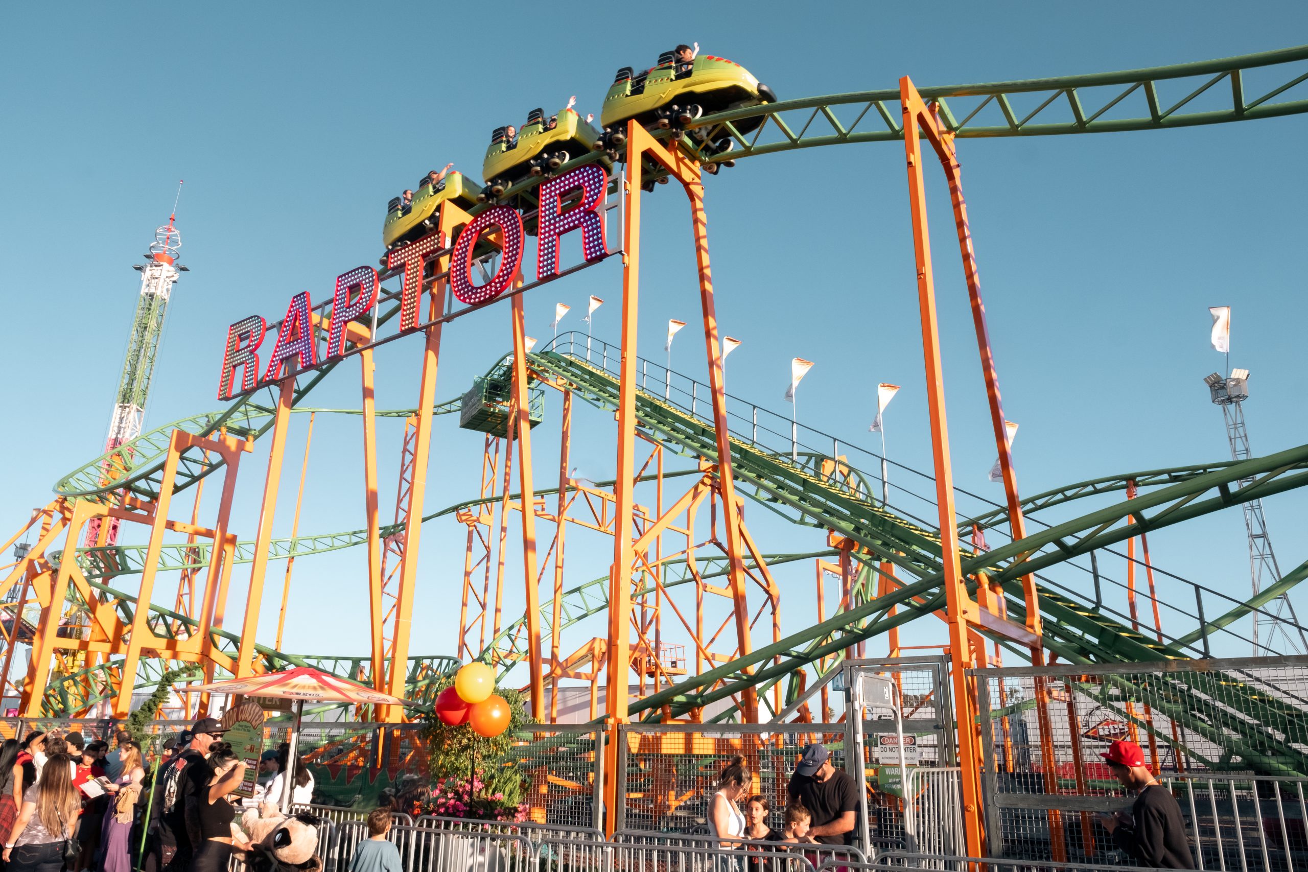 People enjoy a roller coaster ride called Raptor at an amusement park. The green and yellow coaster is elevated, with riders visible at the top, while crowds gather below under a clear blue sky—perfect for a Costa Mesa travel blog snapshot.