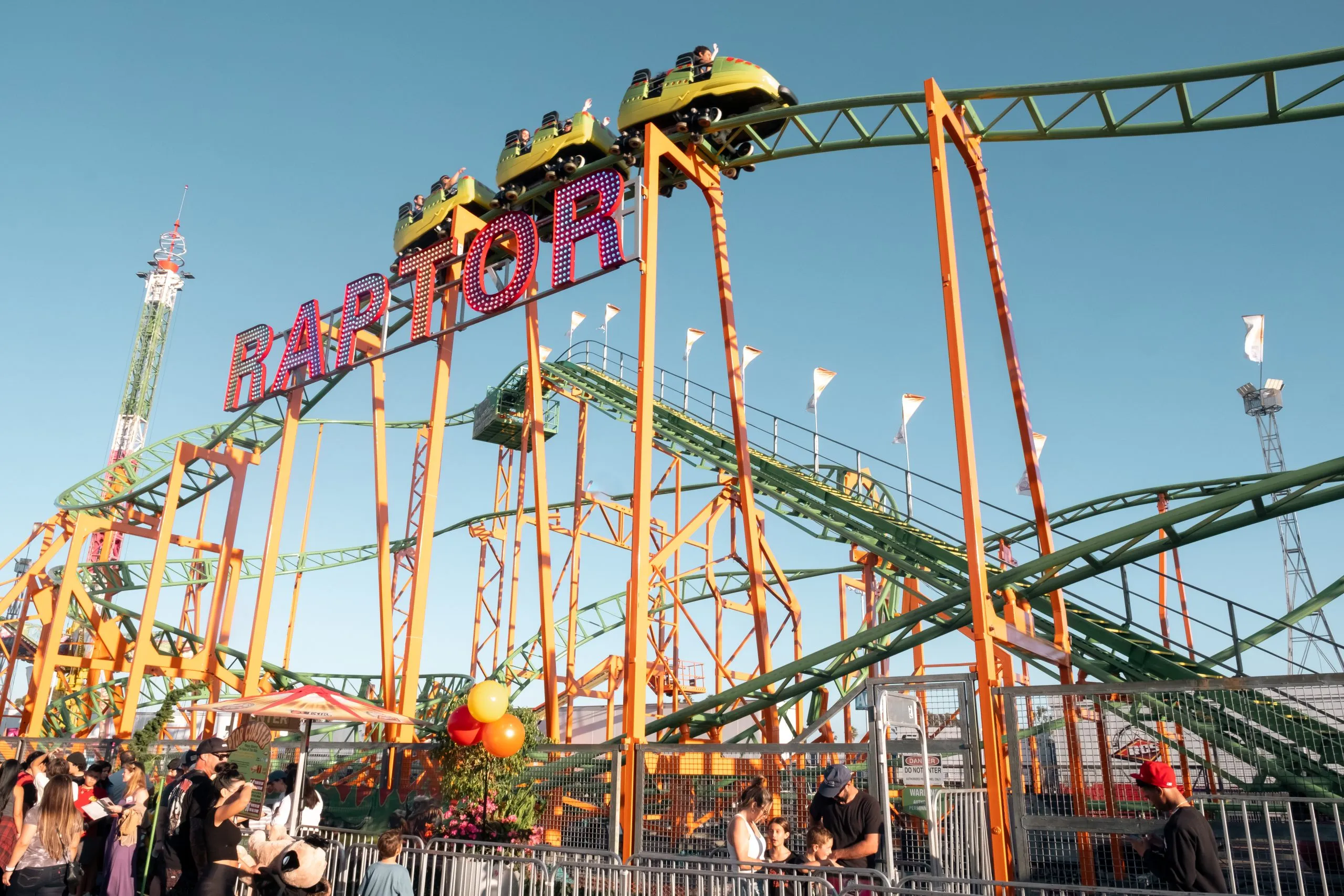 People enjoy a roller coaster ride called Raptor at an amusement park. The green and yellow coaster is elevated, with riders visible at the top, while crowds gather below under a clear blue sky—perfect for a Costa Mesa travel blog snapshot.