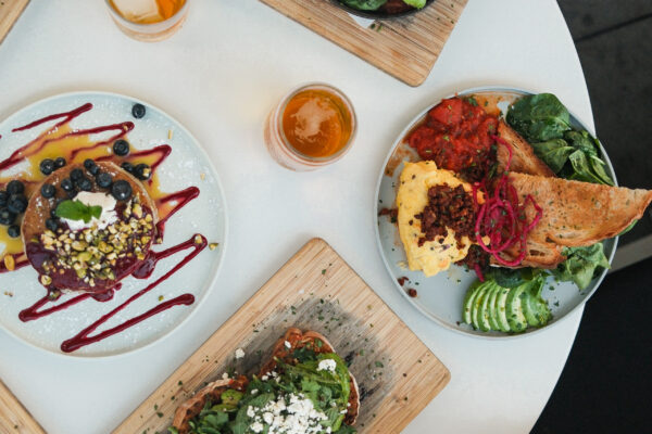 A round white table at Outpost Kitchen Costa Mesa is set with plates of colorful brunch foods—avocado toast, waffles with berries, leafy salads, toast, and drinks—as multiple hands reach in with utensils and glasses.