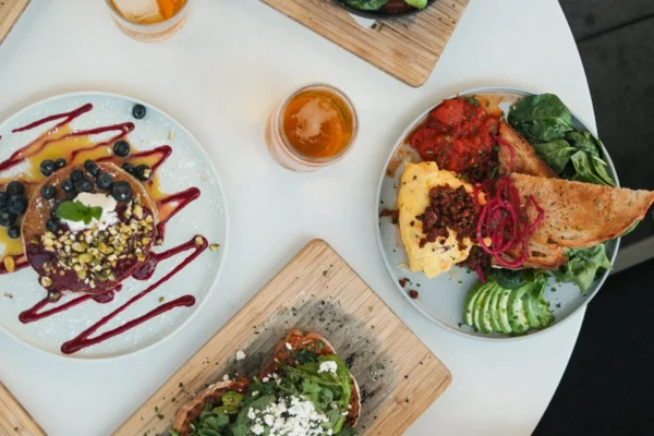 A round white table at Outpost Kitchen Costa Mesa is set with plates of colorful brunch foods—avocado toast, waffles with berries, leafy salads, toast, and drinks—as multiple hands reach in with utensils and glasses.