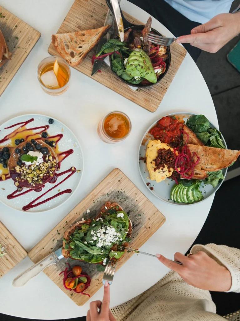 A round white table at Outpost Kitchen Costa Mesa is set with plates of colorful brunch foods—avocado toast, waffles with berries, leafy salads, toast, and drinks—as multiple hands reach in with utensils and glasses.