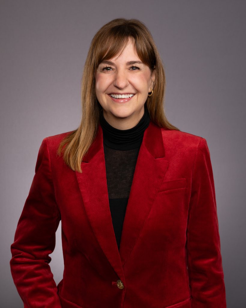 A woman with straight brown hair, wearing a red blazer over a black turtleneck, smiles at the camera against a plain gray background.