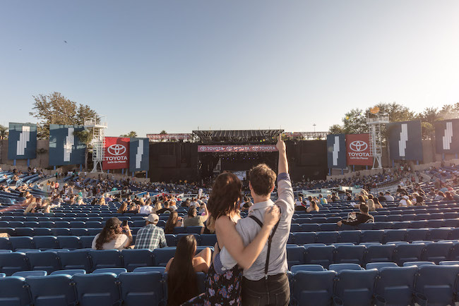 A couple stands in the outdoor amphitheater at the OC Fair Costa Mesa, the woman’s arm around the man as he points toward the stage. Blue seats surround them, with people scattered throughout, waiting for a concert to begin.