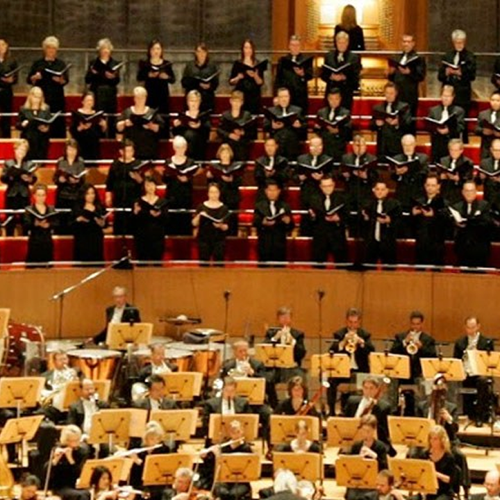 A large orchestra is seated in front of a choir standing on risers in Costa Mesa. The choir members, wearing black attire and holding sheet music, perform in a grand concert hall with red seats and golden accents.