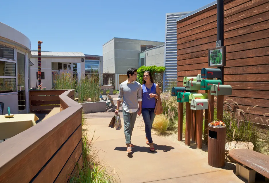 A couple walks together outside on a sunny day, holding hands and shopping bags, passing colorful mailboxes and wooden fences in a modern outdoor shopping area.