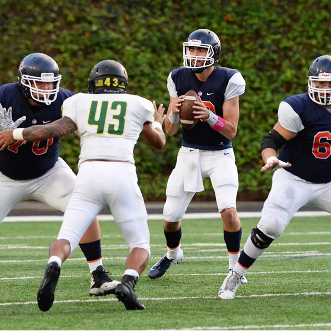 A football quarterback in a dark uniform holds the ball, preparing to pass as teammates block opposing players in white and green uniforms during lively sports events in Costa Mesa, Orange County.