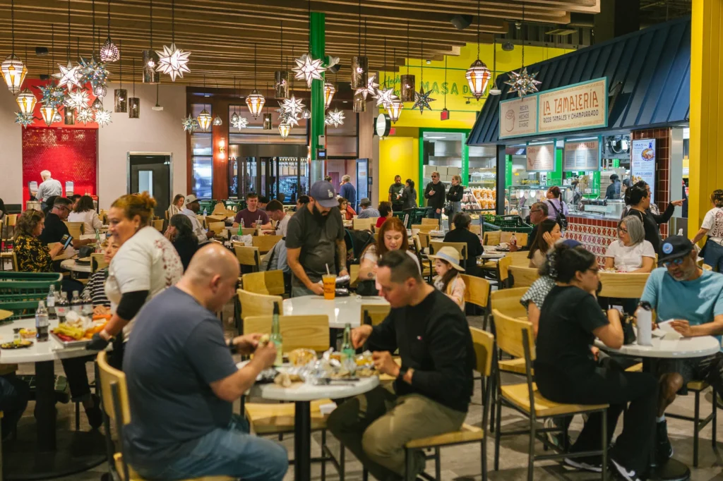 A busy indoor food court in Costa Mesa with people sitting at tables eating and chatting under hanging star-shaped lights, near food vendor stalls with colorful signs—perfect for a Costa Mesa travel blog feature.