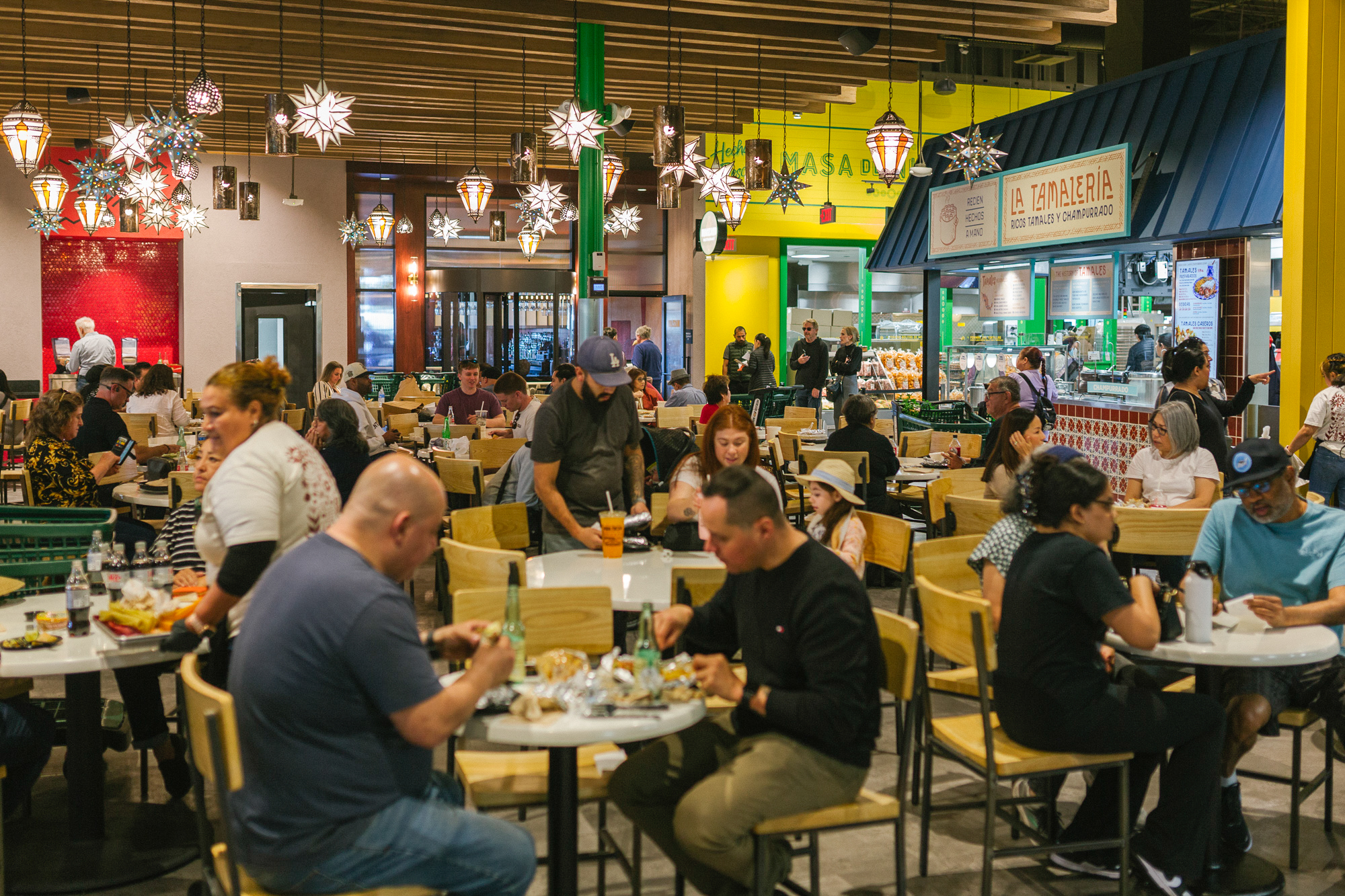 A busy indoor food court in Costa Mesa with people sitting at tables eating and chatting under hanging star-shaped lights, near food vendor stalls with colorful signs—perfect for a Costa Mesa travel blog feature.