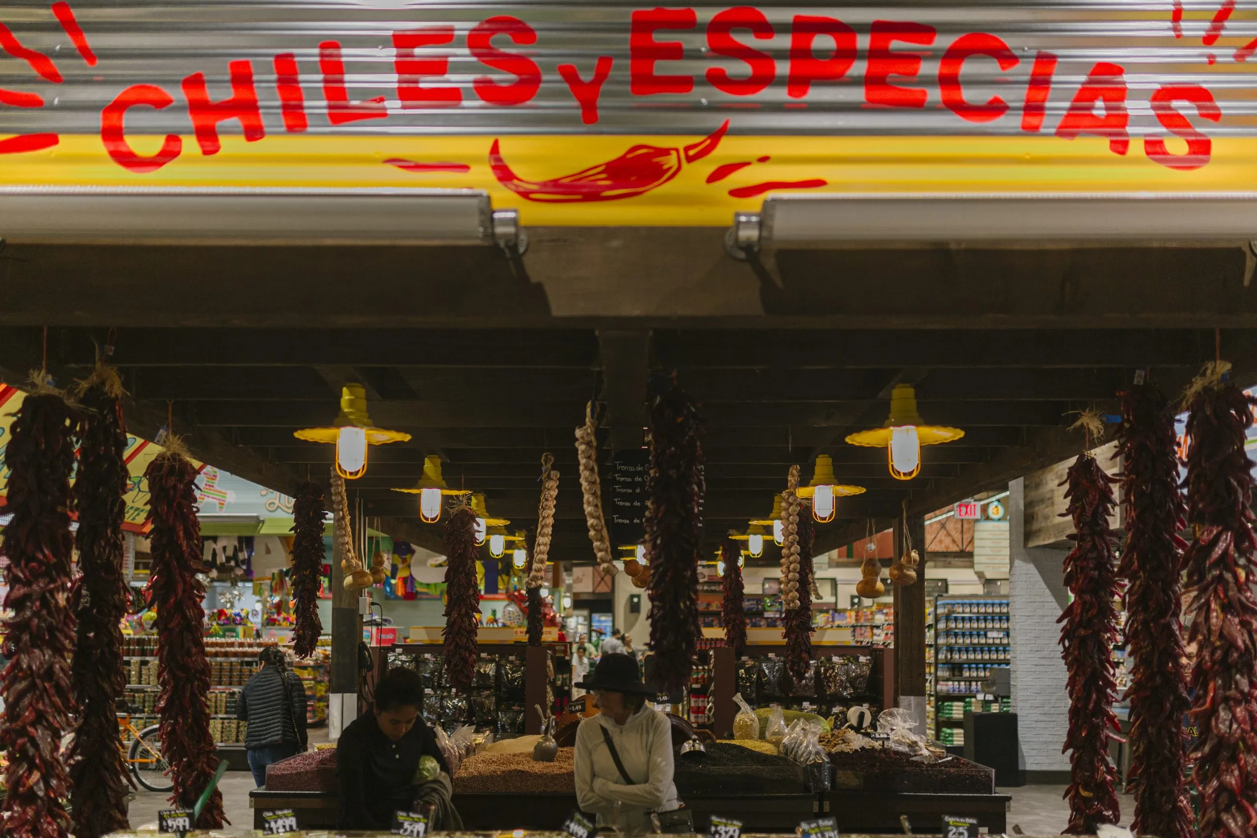 A market stall labeled Chiles y Especias displays hanging dried chiles and spices, with two people standing behind the counter under warm lights—a vibrant scene perfect for any Costa Mesa travel blog.