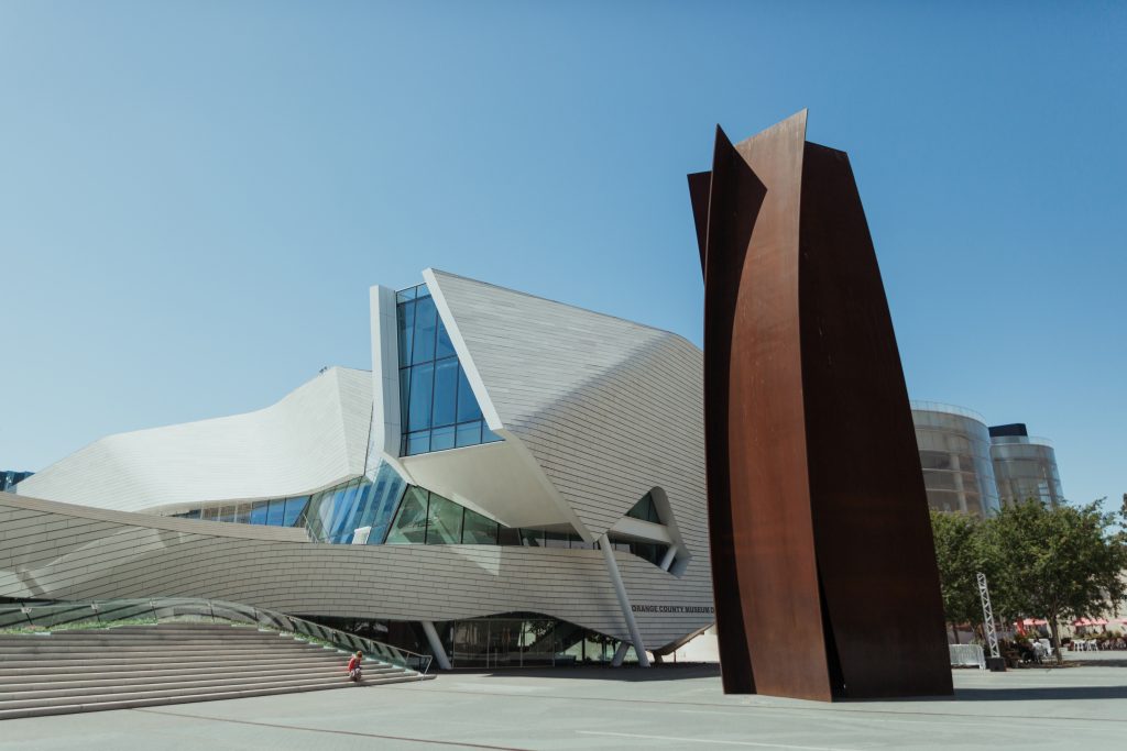A modern building with angular, white, and glass architecture stands beside a tall, abstract rust-colored metal sculpture under a clear blue sky. Trees and other structures are visible in the background.