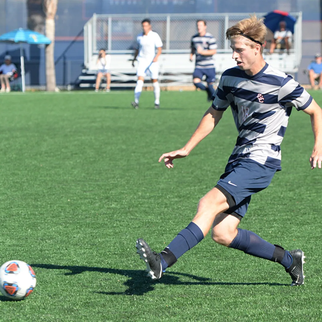 A soccer player in a navy and gray striped uniform kicks a ball on a green field during sports events in Costa Mesa, with teammates and spectators visible in the background.