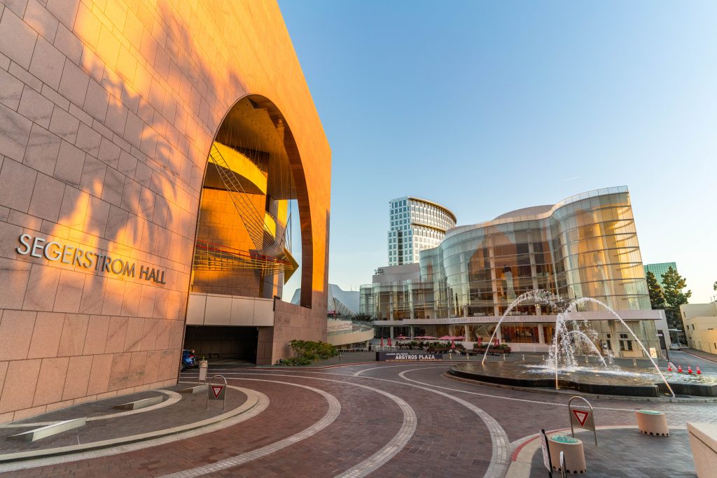 Segerstrom Hall’s curved, golden facade and glass-enclosed lobby glow at sunset. A round fountain with arcing water jets sits out front, and modern buildings rise in the background.