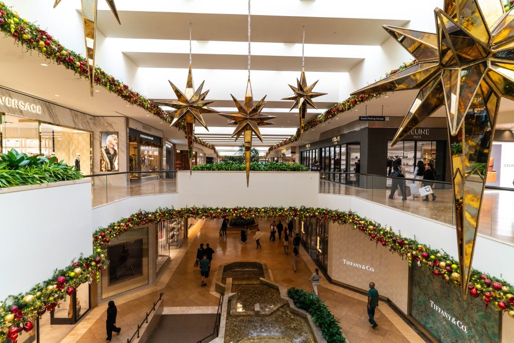 A shopping mall decorated for the holidays with gold star ornaments and garlands. Shoppers walk on two levels, with luxury stores like Versace and Tiffany & Co. visible. A small indoor stream runs on the lower level.