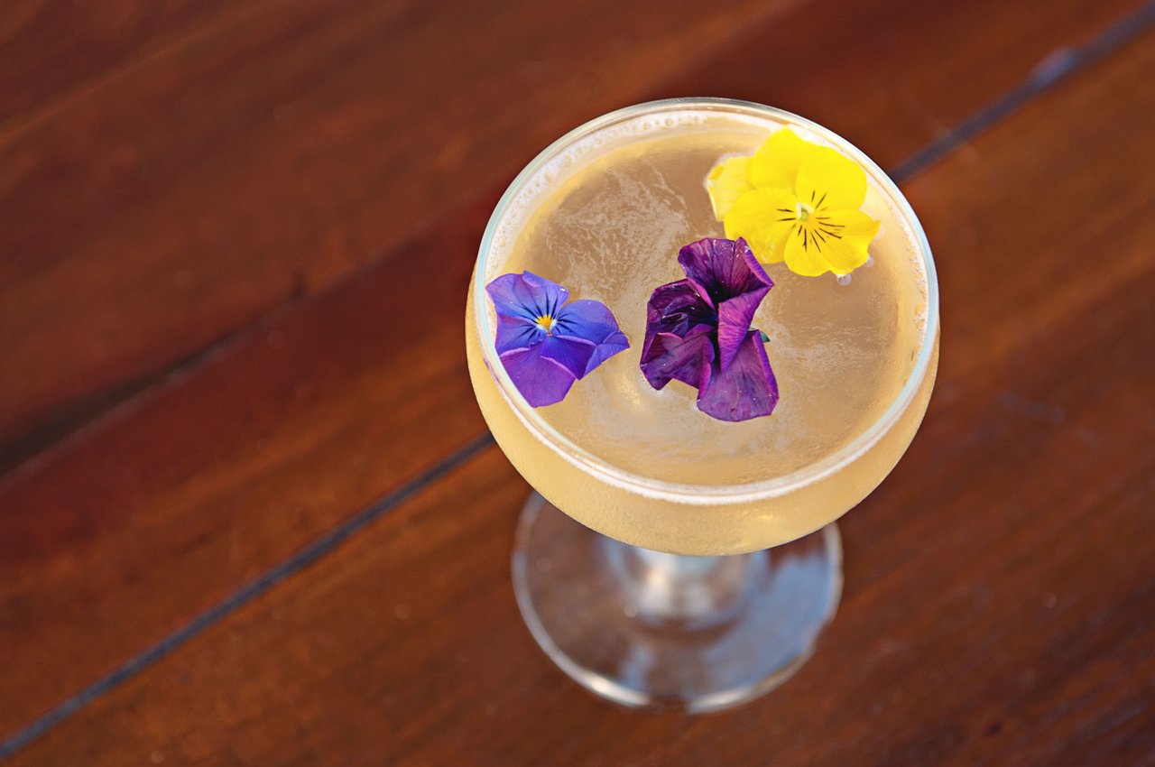 A cocktail in a coupe glass garnished with three colorful edible flowers—yellow, purple, and blue—sits on a wooden surface.
