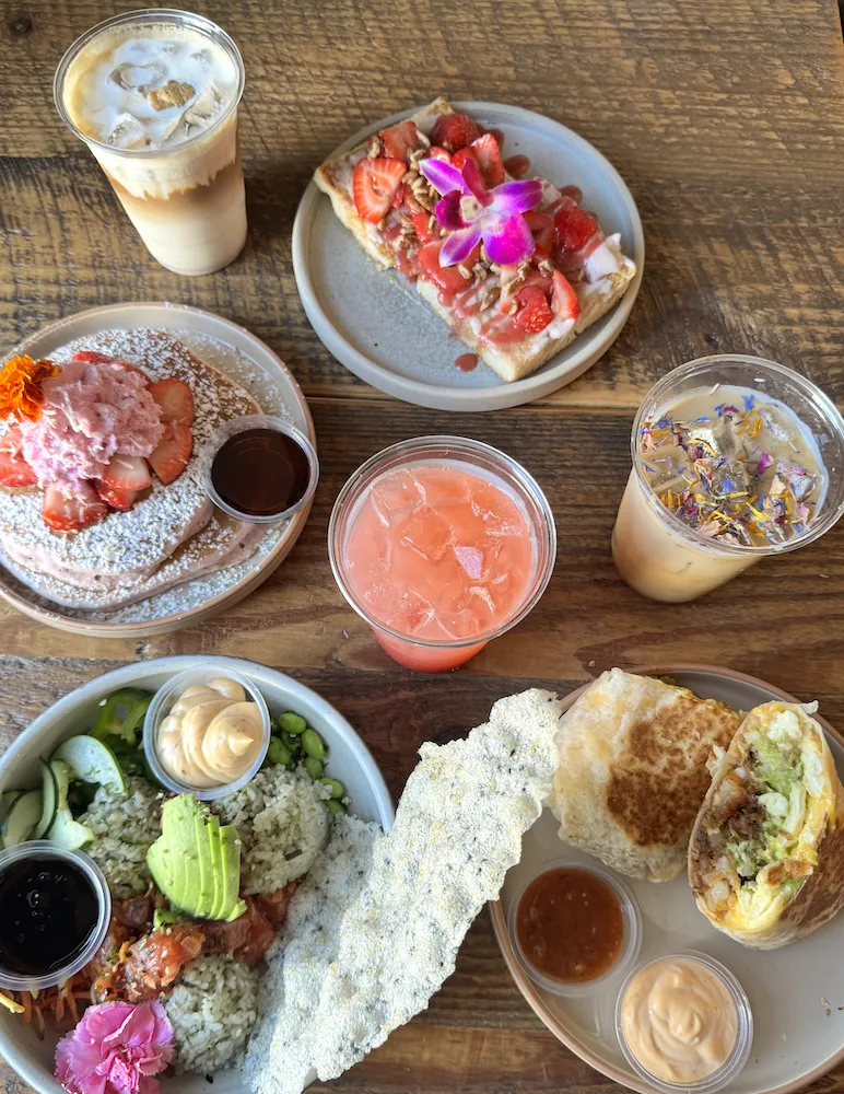 A wooden table with assorted brunch foods: iced coffee drinks, a pink lemonade, a stack of pancakes with strawberries, avocado salad with dressing, a burrito cut in half, and toast topped with strawberries and edible flowers.